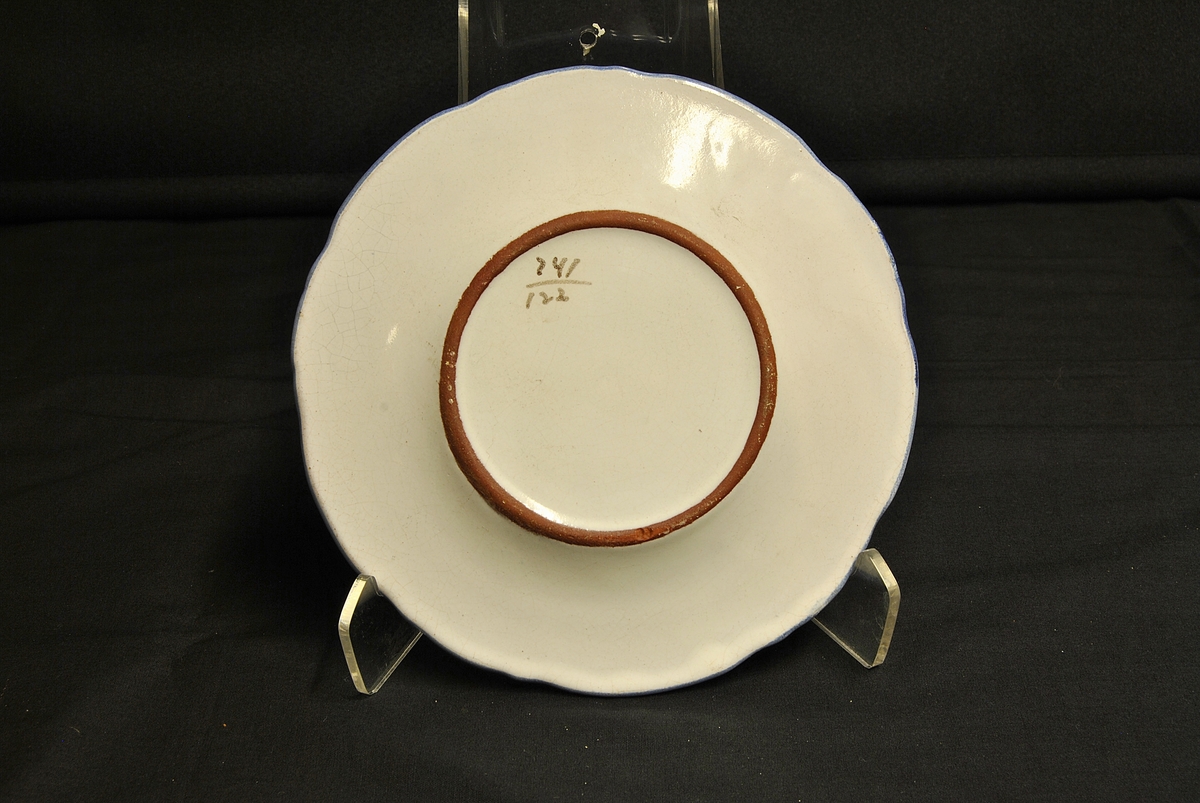 Sirkelforma, kvit tallerken med bølga kant og blomsterdekor: eit stort motiv på midten og fem mindre motiv rundt kanten.
