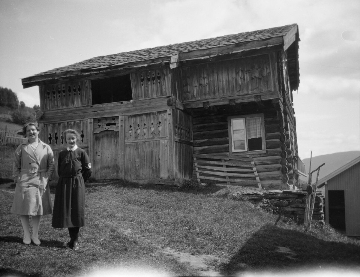 Kvinne i mørk Nasjonalforeningens sykepleieruniform står sammen med en kvinne på tunet forran et gammelt tømmerhus