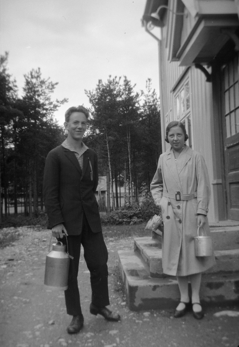 Fra venstre står Ivar Kluften sammen med en kvinne, begge med melkespann i hånden, ved trappen inn til butikken i Furuly, Vålebru.