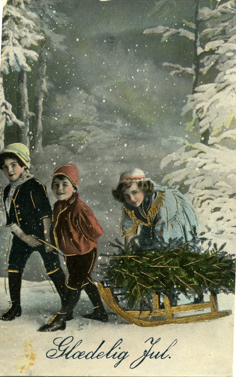 Julekort. Jule-og nyttårshilsen. Vinterlandskap. Tre barn drar et juletre på en kjelke. Barnas klesdrakter er pyntet med gulldekor. Stemplet 23.12.1928.