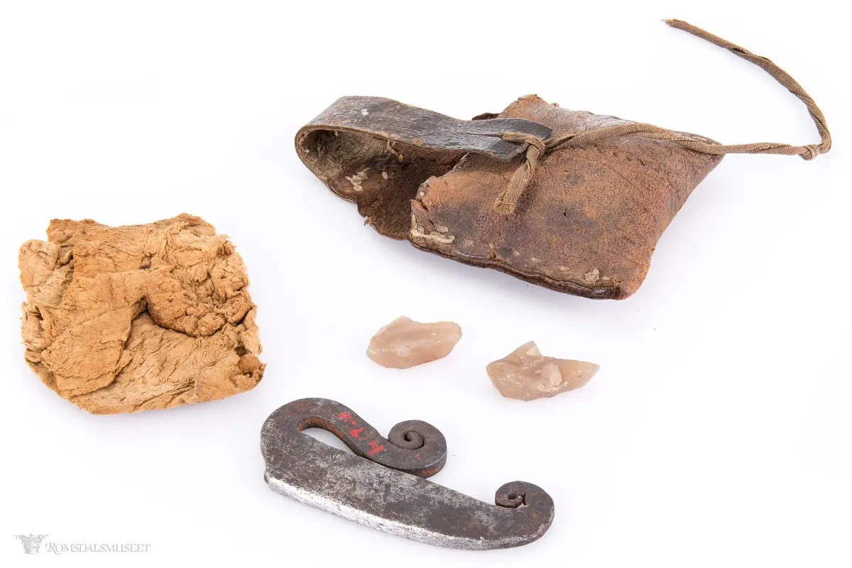 Fyrtøy bestående av en liten lærpose, slagstål, flint og knuskstykker. I posen er det to knuskstykker. Slagstålet har nærmest strykejernsform.