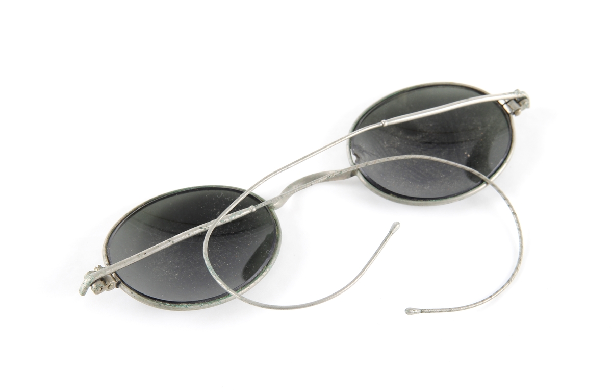 Solbrille med ovale og mørk grå glass. Tynn metallinnfatning med hengsler og to stenger.