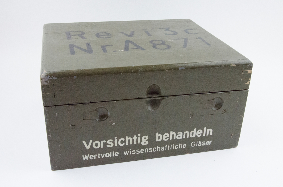 Reflexsikte m/37. Siktet ligger i trälåda som är grön. 
På lådan är det skrivet med svart text på locket och vit text på framsidan.