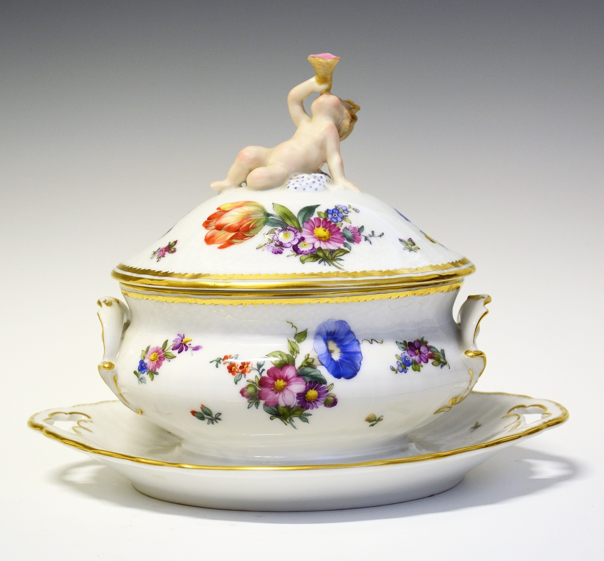 Sauseterrin av porselen dekrorert med håndmalt blomsterdekor og modellert figur av et barn med blåseintstrument som lokknapp. Hanker på hver siden med skjellform. Alle kanter er markert med gull.