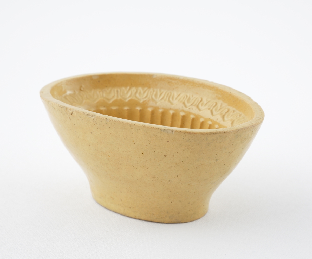 Aladåbform i keramik. 
Oval modell med räfflade kanter, överst en palmettbård. I botten en inpräglad druvklase.
Honungsgul glasyr.