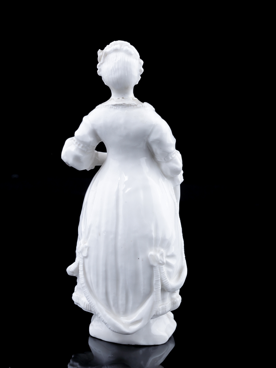 Figurin i vitt porslin. 
Kvinna klädd i 1700-talskläder med kappa. Mycket otydlig signering i botten. 
Troligen Marieberg, möjligen Rörstrand.