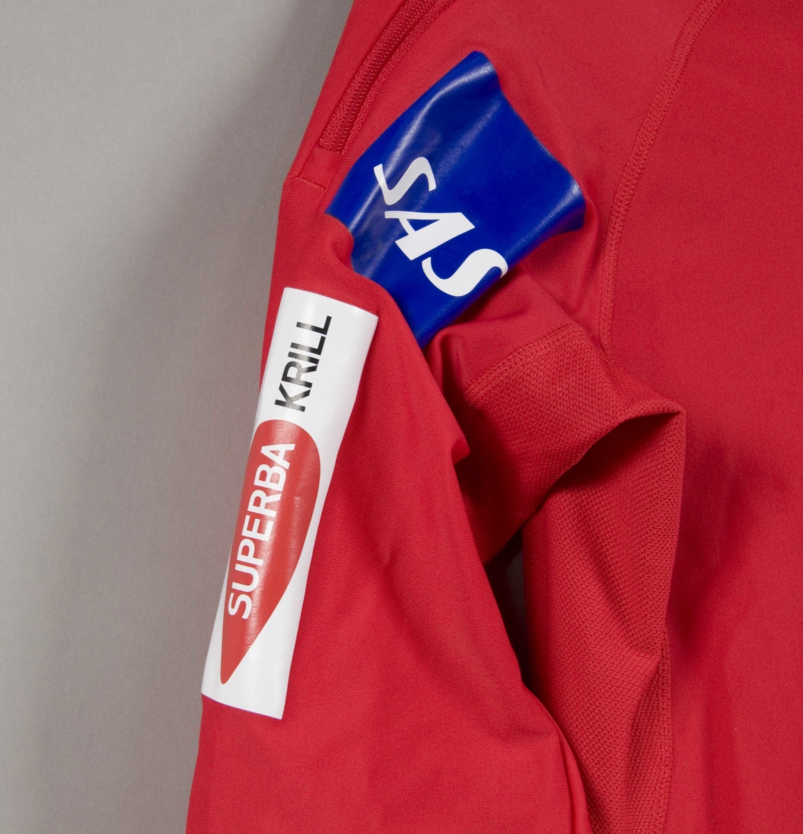 Rød langrennsdress med hvite og blåe striper. På dressen er det flere laminerte logoer for forskjellige sponsorer.