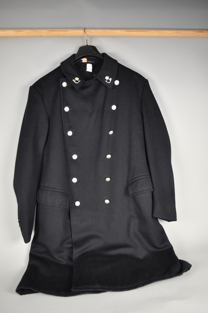 Lang uniformsfrakk med spensel bak. Frakken er sort med sølvfargede postlogomerker og knapper.