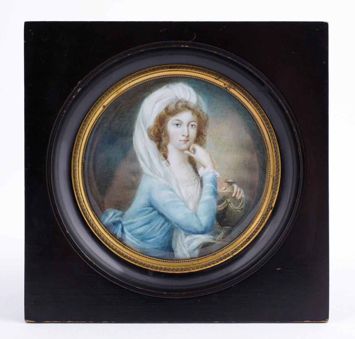 Miniatyrmålning. 
Rund akvarellmålning av kvinna i blå klänning med pärlhalsband och turbanliknande mössa med slöja, som håller om en urna med girlanddekor. 
Bröstbild, halvprofil.
