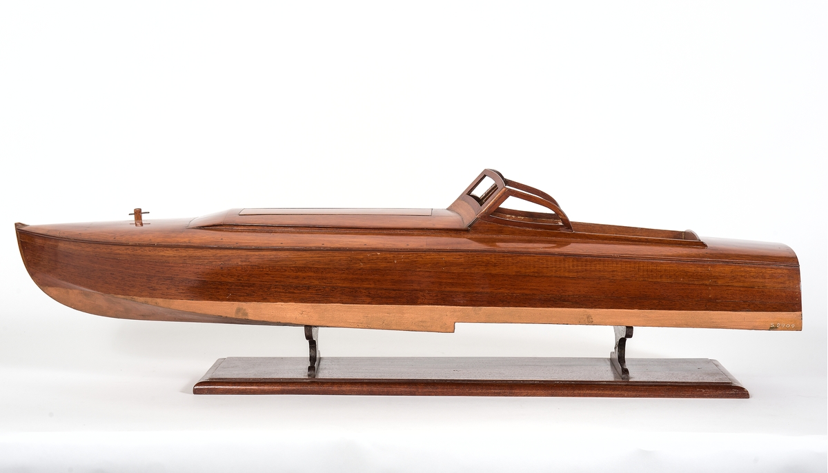 Båtmodell av motoryacht byggd av mahogny på ekspant, halvdäckad. Maskinrum med två luckor, styrbrunn och sittrum. Flat, bronserad botten.
Skrå av mahogny.