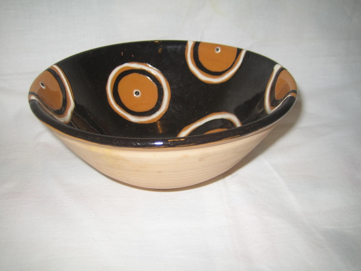 Rund skål i naturfärgad keramik på utsidan och svart glasyr på insidan, dekorerad med cirklar i vitt och svart med en vitsvart prick i mitten.