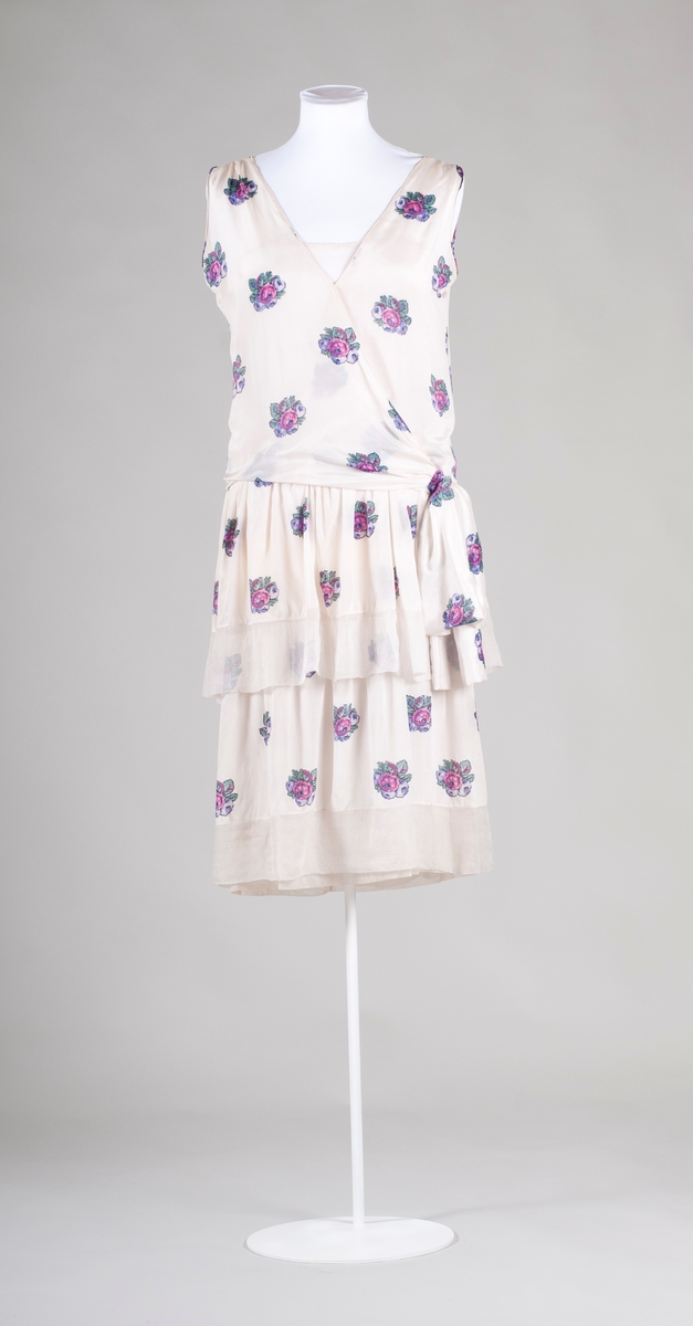 Klänning av vit siden med blomtryck. Kjol med volang och underklänning av tyll. Framstycket går omlott och avslutas med rosett i sidan.  V-formad halslinning. Ärmlös. 1920-tal.
