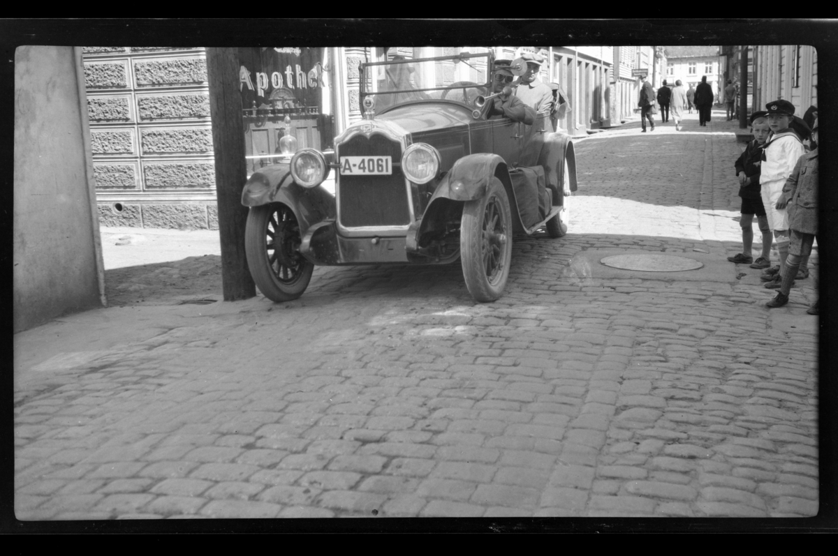 Rolf Sundt Sr. kjører sin Buick gjennom ukjent gate. En gruppe smågutter er tilskuere. Finn Sundt er passasjer. Fotografert 1928.
