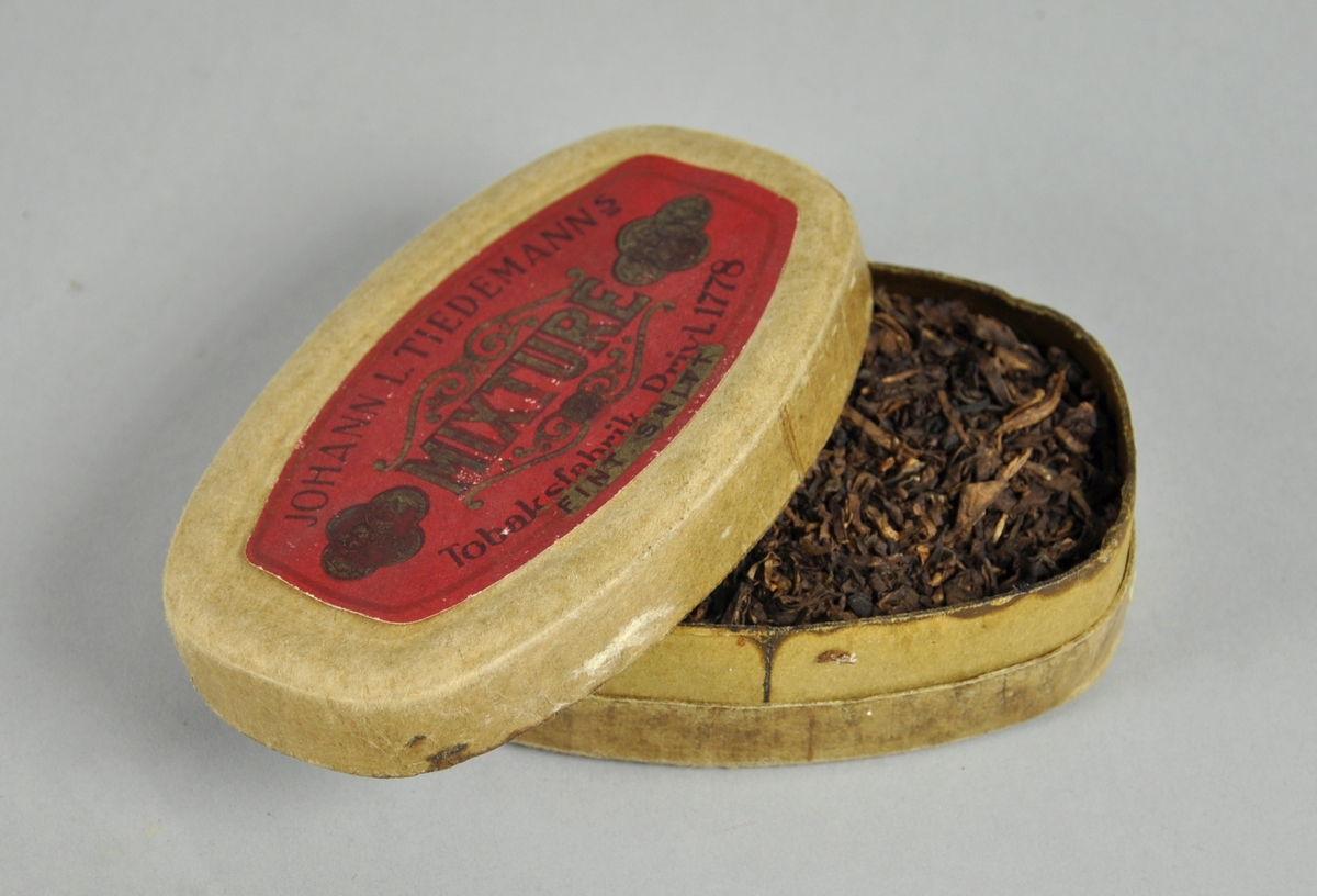Lysebrun, oval tobakkseske av kartong med rød etikett på lokket, og pålimt remse på undersiden med opplysning om pris og vekt. Esken inneholder tobakk.