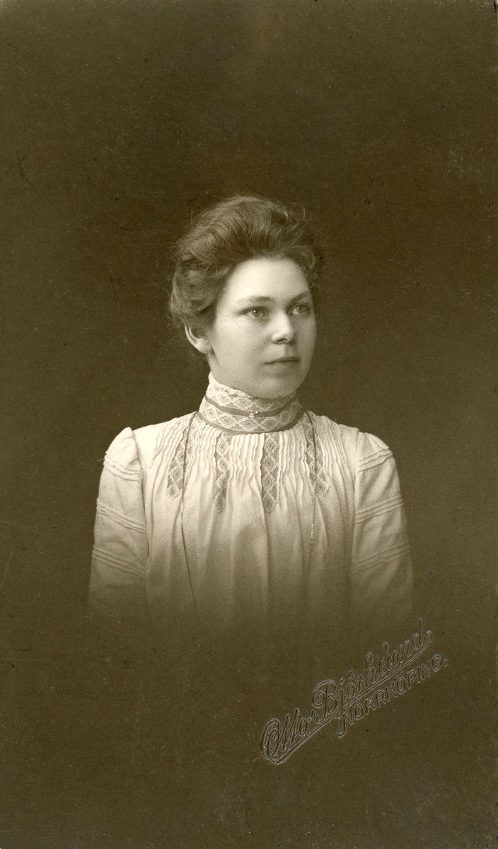 Porträtt av Hedvig Ahlvin omkring 1905. Hon föddes i Söderköping 1878. Födelseattesten uppger att inte bara fadern, utan även modern önskade vara okända. Hur hennes uppväxt fortskred av det skälet är inte känt. Hon påträffas i källorna som inflyttad till Hedvigs församling i Norrköping 1888. Under 1890-talets första del listas hon under rubriken "utom staden boende eller med okänd vistelseort". Med tiden stabiliserades hennes tillvaro. Fotografiet ingår i hennes bevarade ansökan om anställning vid Axel Karlssons manufakturrörelse i Linköping. Rimligtvis erhöll hon platsen. Under alla omständigheter inflyttade hon till Linköping 1906 och uppges vara butiksbiträde.