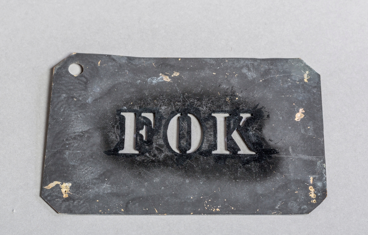 Rektangulær sjablong i metall for maling og merking, med ordet FOK.

Den ene siden av sjablongen er malt svart.

Flekker av svart maling fra merking på sjablongen.