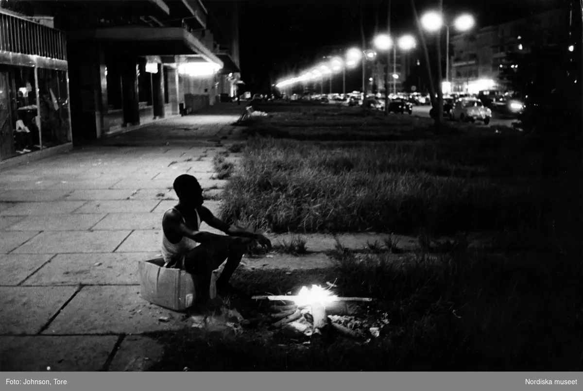 Kongo. En nattväktare sitter på en kartong och har tänt en brasa på gatan utanför en belgisk butik på Boulevard Albert 1er, Leopoldville.