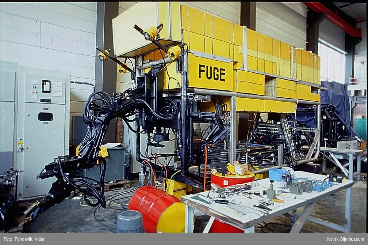 En stor ROV med en robotarm og lyskastere foran står plassert i en verkstedhall.