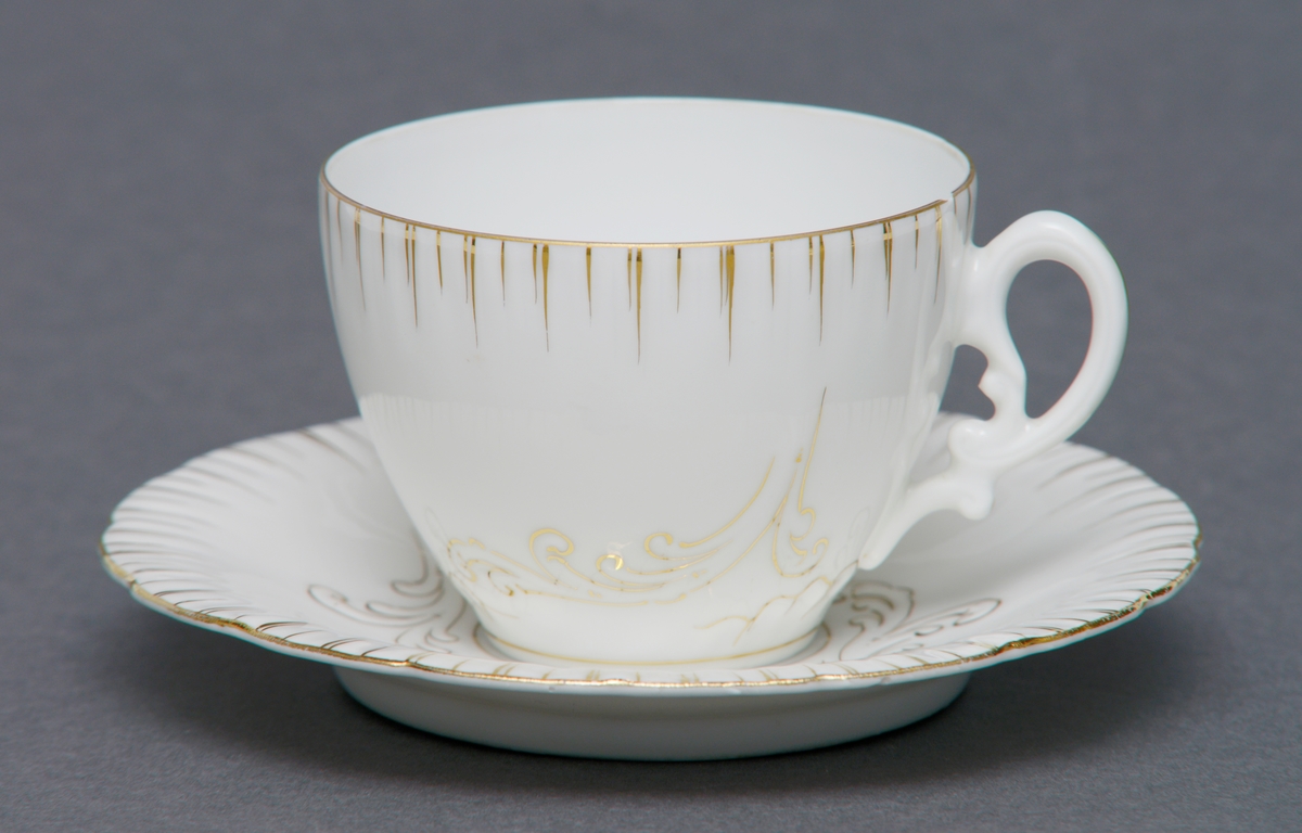 Sirkulært tefat i porselen med glasur. Dekorert med håndmalt gullforgylninger over relieff i porselenet. Bølgete kant. Tilhørende kopp (se relaterte objekter).