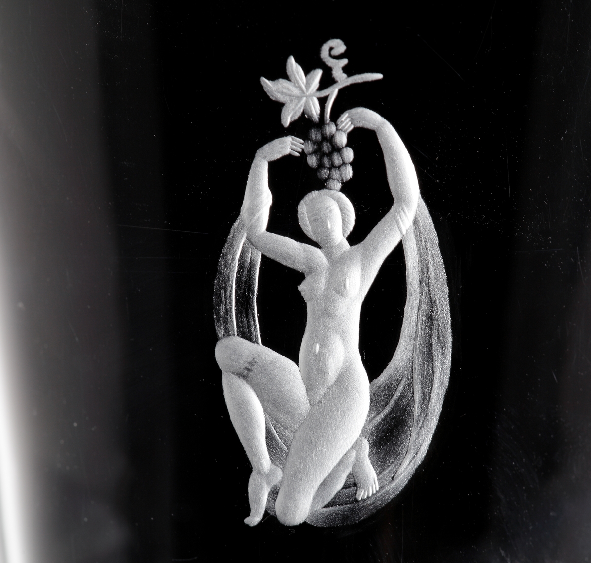 Trumpetformad vas med klack. Graverad med figurmotiv, mattslipad mynningskant samt matt olivslipad dekor på nedre del av vasen. 
Gravör Karl Rössler, verksam vid Orrefors glasbruk 1923-66.