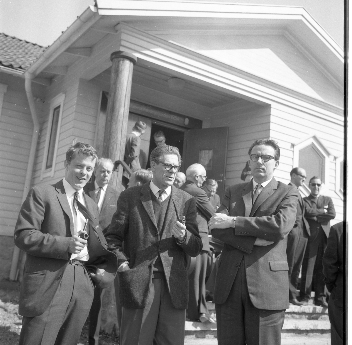H Hall-konferens i Lövhult 1964. Okända kostymklädda piprökande män utanför en byggnad samlade i olika små grupper.
