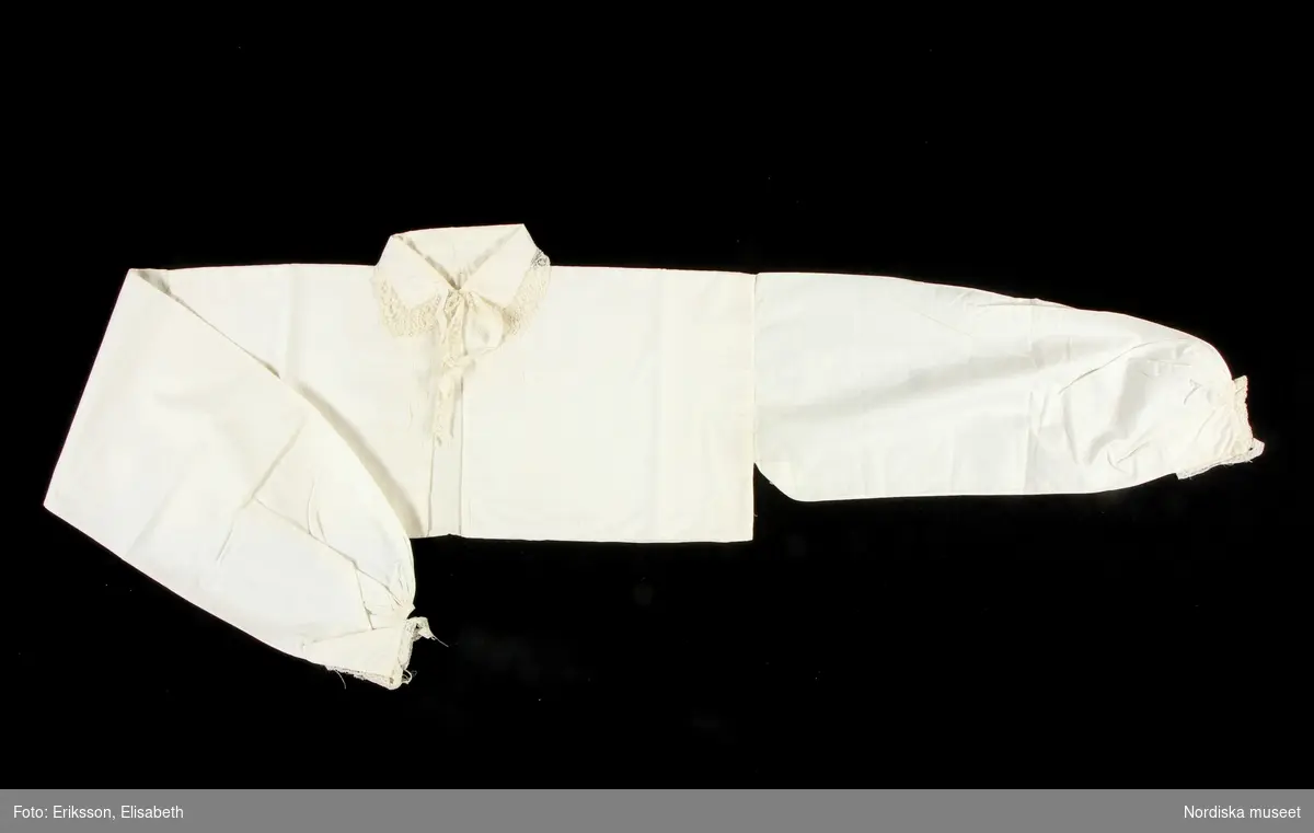 Huvudliggaren:
"a-r Kvinnodräkt. a. kjol, b. förkläde, c. kappa, d. tröja, e. överdel
f.1 - f.4 huvudbonad, (4 delar), g. bälte, h. kjolsäck, i. handskar, j: silkesduk, k. bokkläde, l. psalmbok, m.1- m.2 Bältehängen med silversked och skedpåse, n. nåletui, o.1 kniv, o.2 gaffel, o.3 slida av saffian, p. lit. påse, q. kedjekniv, r. ring.  Ink. 13/7 1903 [från] Eriksson, Erik, herr, Kåsta, Vingåker jämte 95.162 [...]. [Brukningsort:] Södermanland Oppunda hd Vingåker sn."

e. Överdel till kvinnodräkt, kort modell som når nedanför brösten,  av vit tunn bomullslärft, ett bålstycke fodrat med vit bomullskypert, öppen fram, rak nedvikt krage med en liten broderad bård i vittbomullsgarn, Både krage och framkanter kantade med en  2,5 cm bred maskinknypplad uddspets. Bitar av maskinspets även som applikation vid sidan om framkanterna. Knäpps i halsen med hake och hyska  samt ett sekundärt ditsytt bomullsband att knyta.. Vidsydd lång ärm  med r spjäll för rörelsefrihet. Ärmen rynkad mot 0,5 cm bred ärmlinning, rynkorna knäppta med fin sicksacksöm, på ärmlinningen liten broderiad ranka, och i kanten smal maskinspets,  ärmsprund med förstärkning, knäppt med liten hake och hyska ,  
/Berit Eldvik 2010-08-27




