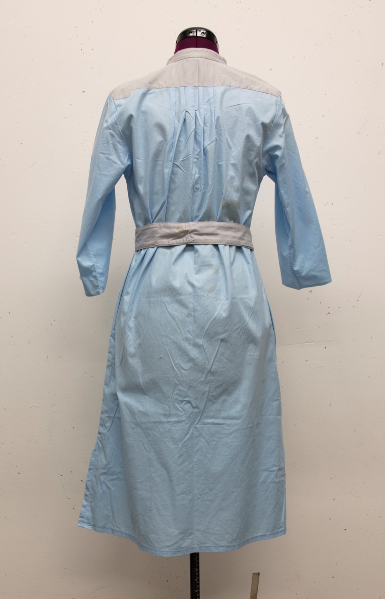 En klänning i blått bomullstyg. Garneringseffekter i gråt tyg. Skärp hör till.
a) klänning
b) skärp