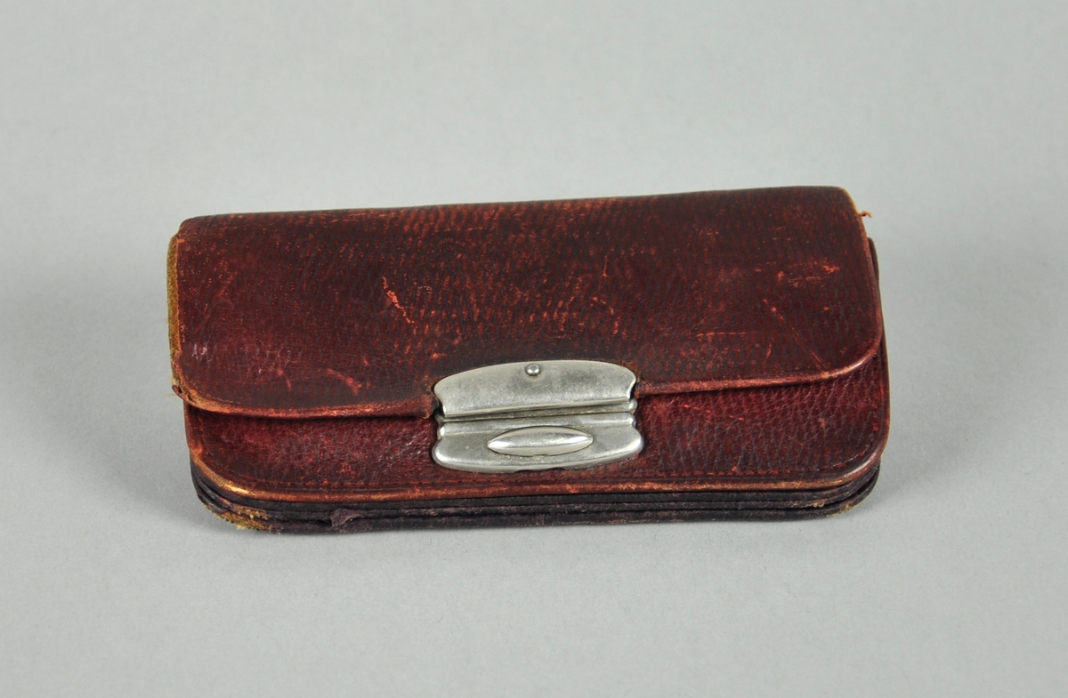 Lommebok av skinn, med lås i metall. Flere lommer kan trekkes ut. En av lommene har metallås.