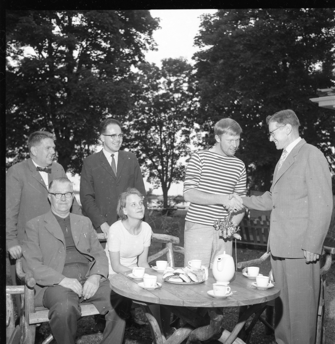 Einar Hafström och okänd flicka sitter vid ett runt bord med kaffekoppar, kaffekanna och bullar. Stående bakom dem från vänster är Sten Roman och Ingmar Lago. Till höger står en ung man i randig tröja med en liten ljuskrona i handen. Han tackar, sannolikt, en kostymklädd äldre man för densamma.