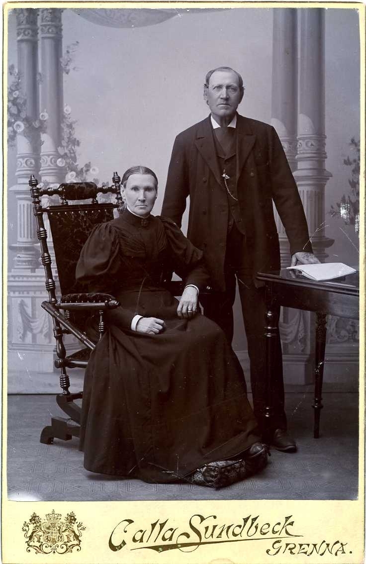 Kabinettsfotografi: Maja-Stina Johannesson sittande i en gungstol med maken Carl stående bredvid, handen vilande på ett bord med uppslagen bok.