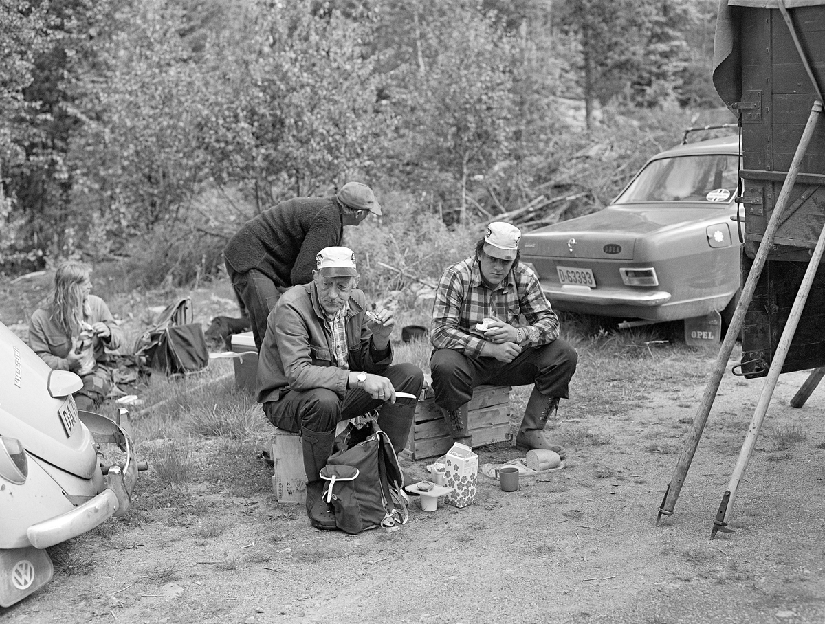 Arne og Arnstein Norsted, far og sønn fra Osen i Hedmark, spiser brødskiver under pause i tømmerfløtinga i elva Søndre Osa, næmrmere bestemt ved Kvernfallet. Fotografiet ble tatt i 1981. Bak Norsted-karene satt Kenth Mokkelbost fra Flisa i Åsnes og skar seg brødskiver mens Nikolai Myrene fra Osen sto bøyd over ryggsekken sin. Helt til høyre i bildet ser vi en lastebil som ble brukt til å trnsportere mat og utstyr nedover langs vassdraget etter hvert som fløterlaget arbeidet seg nedover mot Rena. Vi ser også bakpartene av to personbiler, ei Folkevognboble og en Opel Kadett.