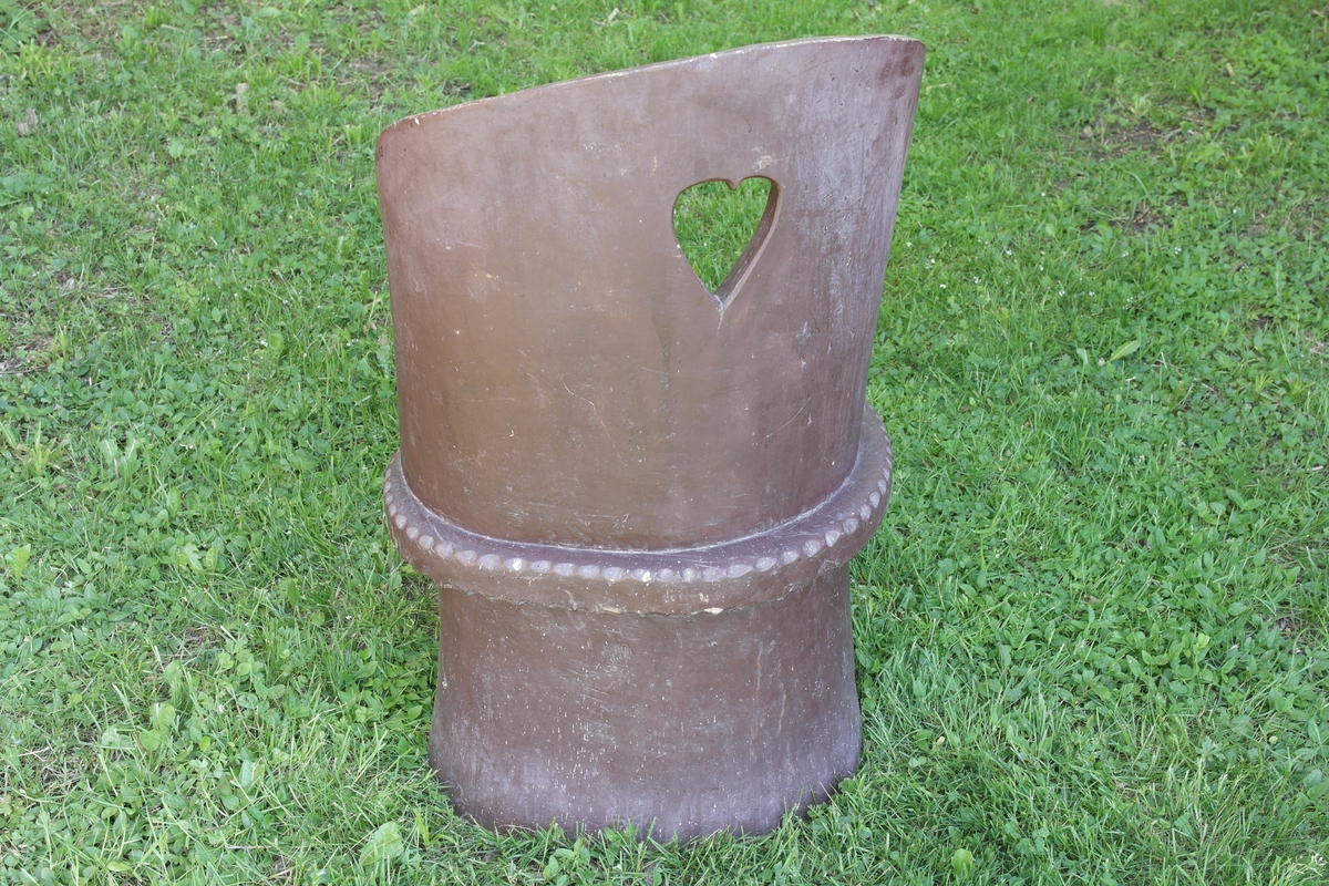 Kubbestol i hugget og skåret osp. Stolen er pusset og malt brun. Understellet og ryggen er skåret i ett stykke. Hjerteformet utsparing i ryggen.