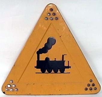 Varningsmärke för vägtrafikanter: Varning för korsande järnväg. Skylten är formad som en trekant, och är tillverkad av aluminiumplåt med reflekterande gul färg. Den visar ett stiliserat svart ånglok i mitten, och har sex stycken glasknoppar i varje hörn.

Stolpfäste på baksidan.