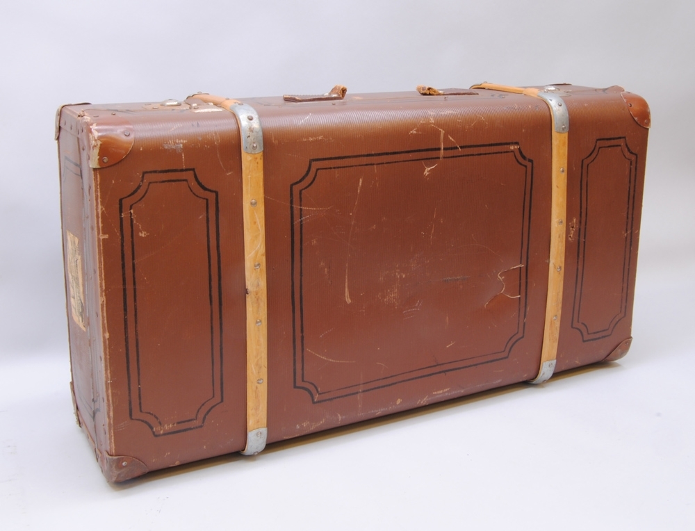 Brun resväska av papp med stomme av trä och två stycken ribbor av böjträ runtom. På väskans sidor är det målat ett dekorativt ramverk av svarta linjer likt speglar på dörrar eller väggpaneler. Resväskan har förstärkningar av metall i hörnen och låsanordning på var sida av handtaget.
Handtaget (:2) är av läder med en separat hållare för adresslapp med adressuppgifter tillhörande väskans ägare (se personuppgifter). På ena kortsidan sitter några lager av klistrade etiketter. På den översta etiketten går det att urskilja vissa ord: "Ankomst", "UMEÅ", "Till", "Över", "snälltåg", "Antal 2", "Tåg 2732". Den handskrivna texten är svår att läsa. På motstående sida sitter också flera lager av etiketter varav den översta är fullständig. Där står det "SJ Snälltåg", "Ankomstdatum:", "Från UMEÅ", Till: Eskilstuna", "Över: Vus.-Bä Sala-Kolbäck", "Antal kolli 3", "435", Totalvikt kg 50.", Tåg 2748", "Datum 4/1 1959". Längst upp i högra hörnet nedanför "Ankomstdatum" är det skrivet på snedden "129" samt "5/1".
Inuti är väskan klädd med mönstrat papper vars prickar och streck bildar ett rutmönster. I botten ligger ett flerfärgat mönstrat vaxat skyddspapper med rosa prickar och linjer. På insidan av locket sitter två gröna lappar som lagningar. Mellan lock och botten sitter bomullsband som håller locket i 90 graders vinkel från botten då väskan är öppen och bomullsband med spännen tvärsöver bottendelen för att hålla väskans innehåll på plats vid transport.