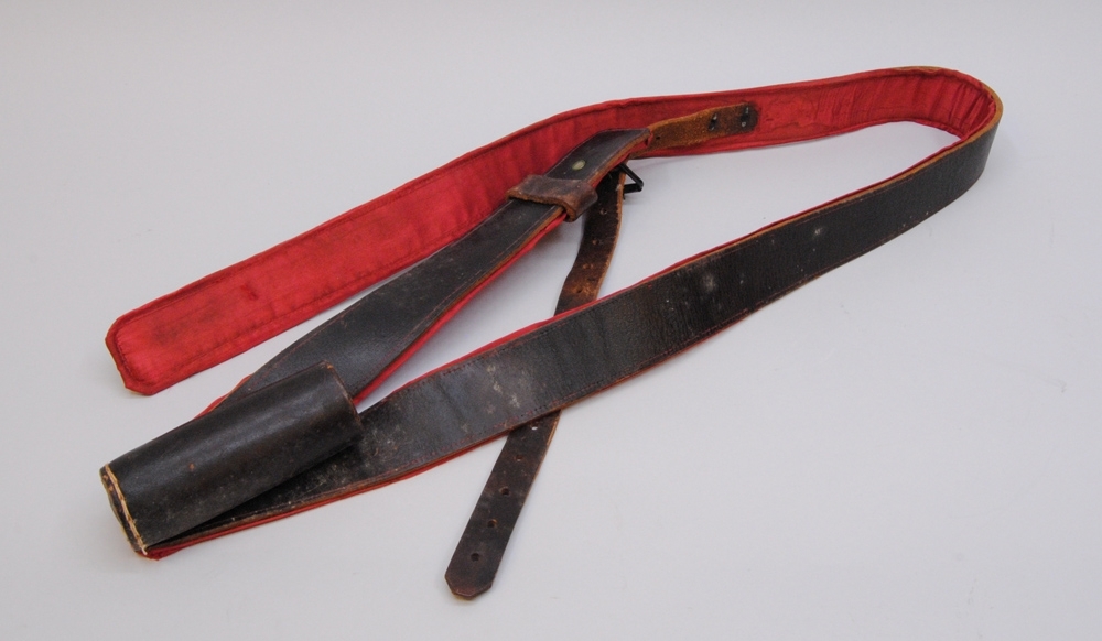 Bandolär, bärrem, av läder med rött sidentyg på insidan och spännen av metall. Tillhör fana Jvm 21169:1.