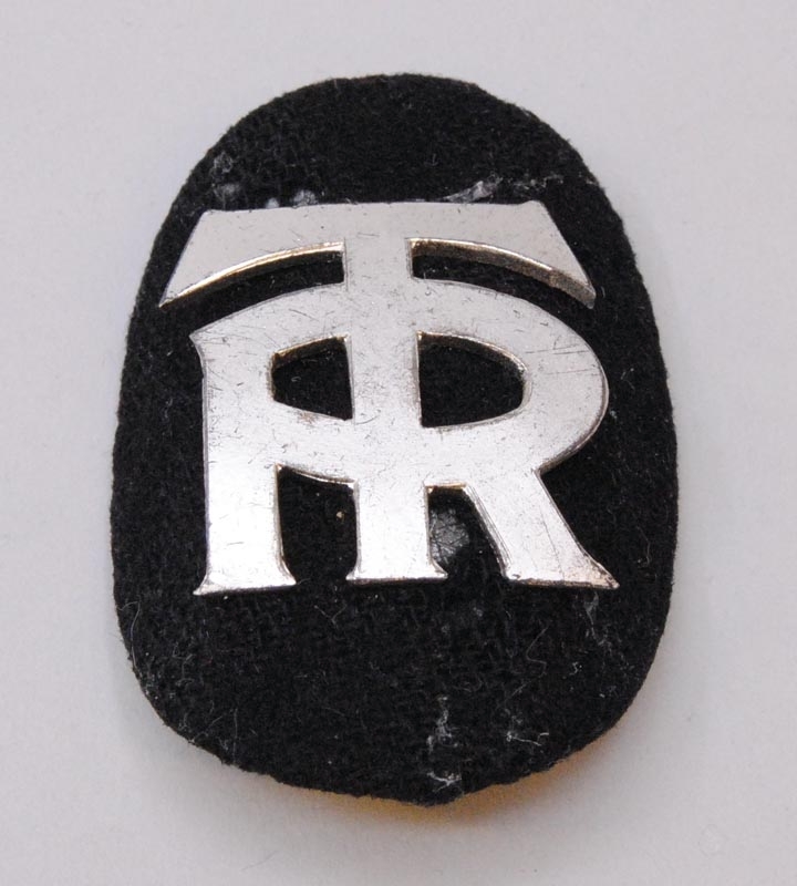 Mössmärke av metall med bokstäverna "TR" sammanlänkade mot en svart bakgrund av textil. Nål på baksidan. Mössmärket har formen av en stående oval med kupad insida.