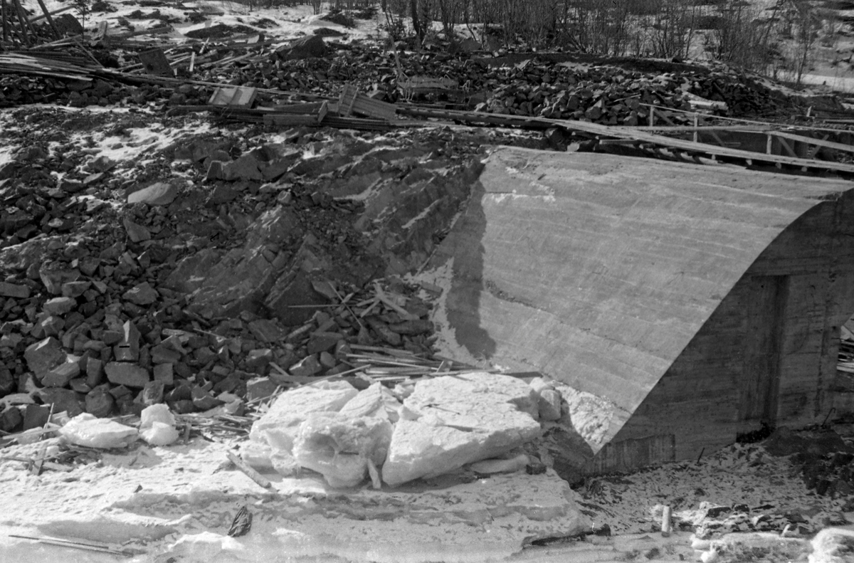 Bygginga av ny inntaksdam ved Eidefoss kraftverk i Ottavassdraget i 1952 (Vågå kommune).  Fotografiet er fra et nesten tørrlagt elveløp gjennom en berghammer.  Damarbeidet ble, typisk nok, utført i overgangsfasen mellom vinter og vår, da vannføringa var minimal.  Stengselet for damåpningen var ennå ikke kommet på plass.  I bakgrunnen skimtes utsprengte masser og en del forskalingsmaterialer.  Det første kraftverket ved Eidefossen ble bygd i 1916-17.  Den nye betongdammen fra 1951-52 ble støpt med henblikk på å utvide magasinkapasiteten, slik at en kunne installere nok et aggregat i kraftstasjonen.