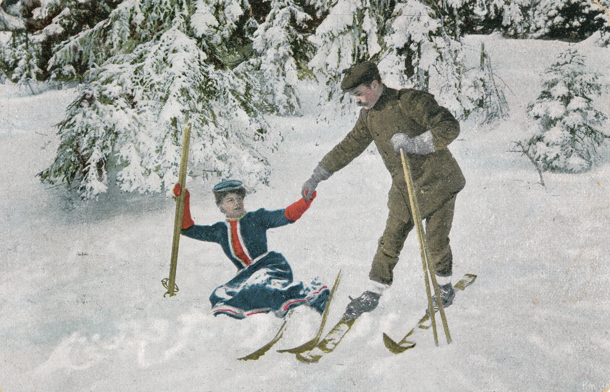 Postkort. Motivet på kortets fremside viser mann og kvinne på skitur i vinterlandskap. Mannen hjelper kvinnen etter at hun har falt i snøen.