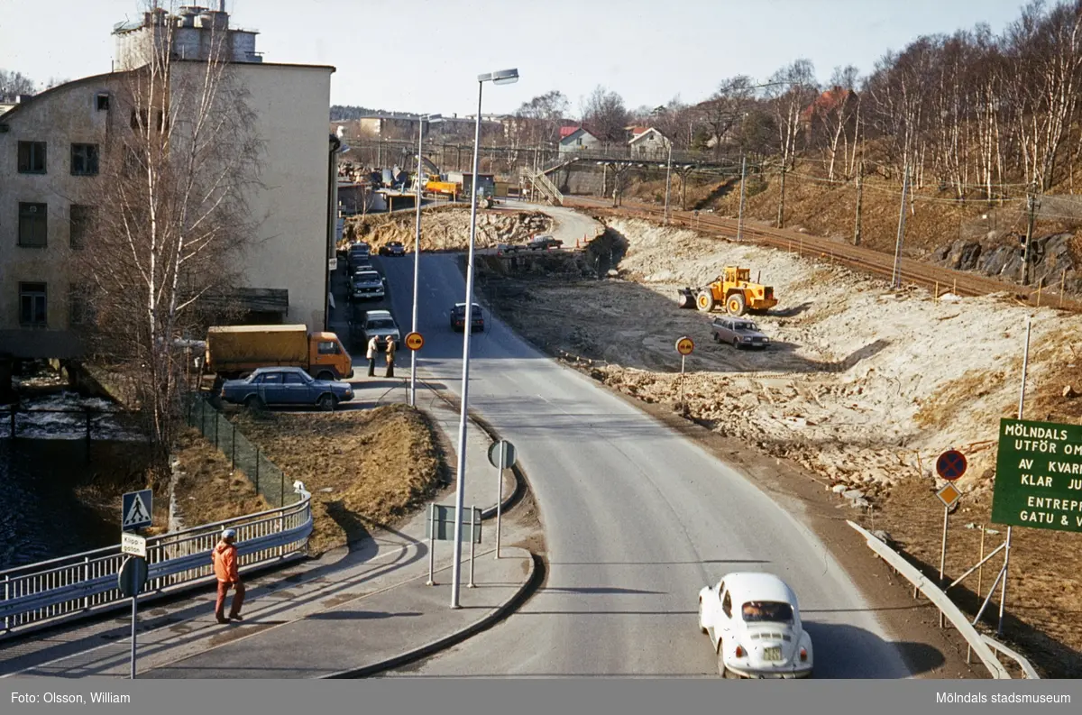 Vy från Stensjögatans bro västerut, okänt årtal. En vit Volkswagen "Bubbla" kör Pixbovägen ner. Till vänster ses ett staket och under Rackarebron rinner forsen. Byggnaden är "Strumpan" (Kvarnbygatan 10-14) där bilar står parkerade utanför. Till höger pågår vägarbete för att flytta Kvarnbygatan. Längst bort löper järnvägen Boråsbanan.