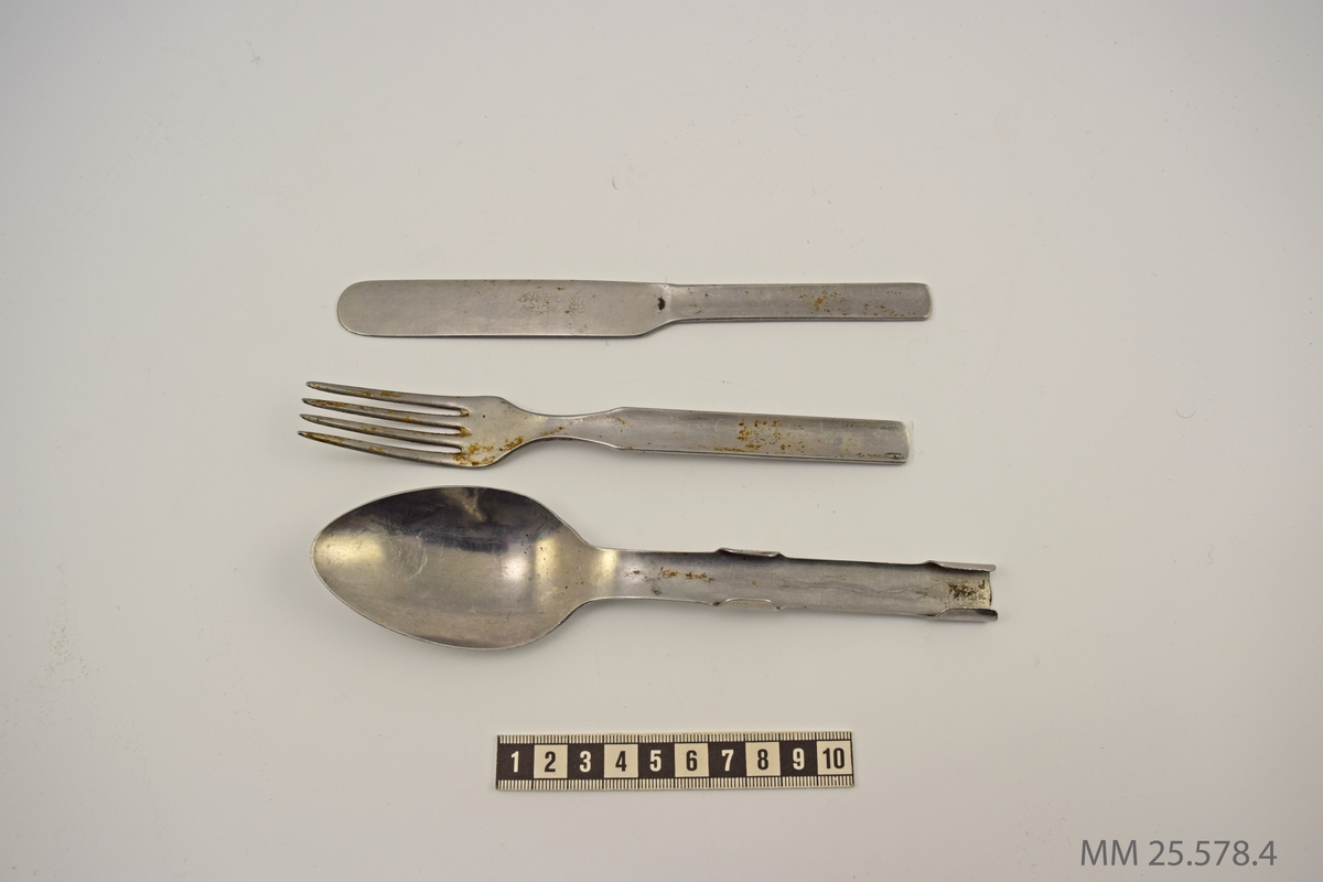 Kniv, gaffel och sked i metall. Skeden har två byglar på skaftet under vilka gaffeln och kniven kan fästas.