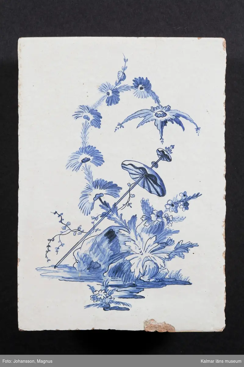 KLM 23946. Kakel. Kakelugnskakel. Ett plant bladkakel med vit/ljusgrå glasyr med rokokomönster i blått, blommande kvist och herdinneparasoll.