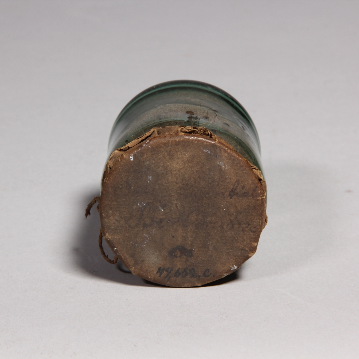 Apoteksburk av grönt glas, cylindrisk med överbundet lock av papper med snöre. Innehåller brunt pulver.