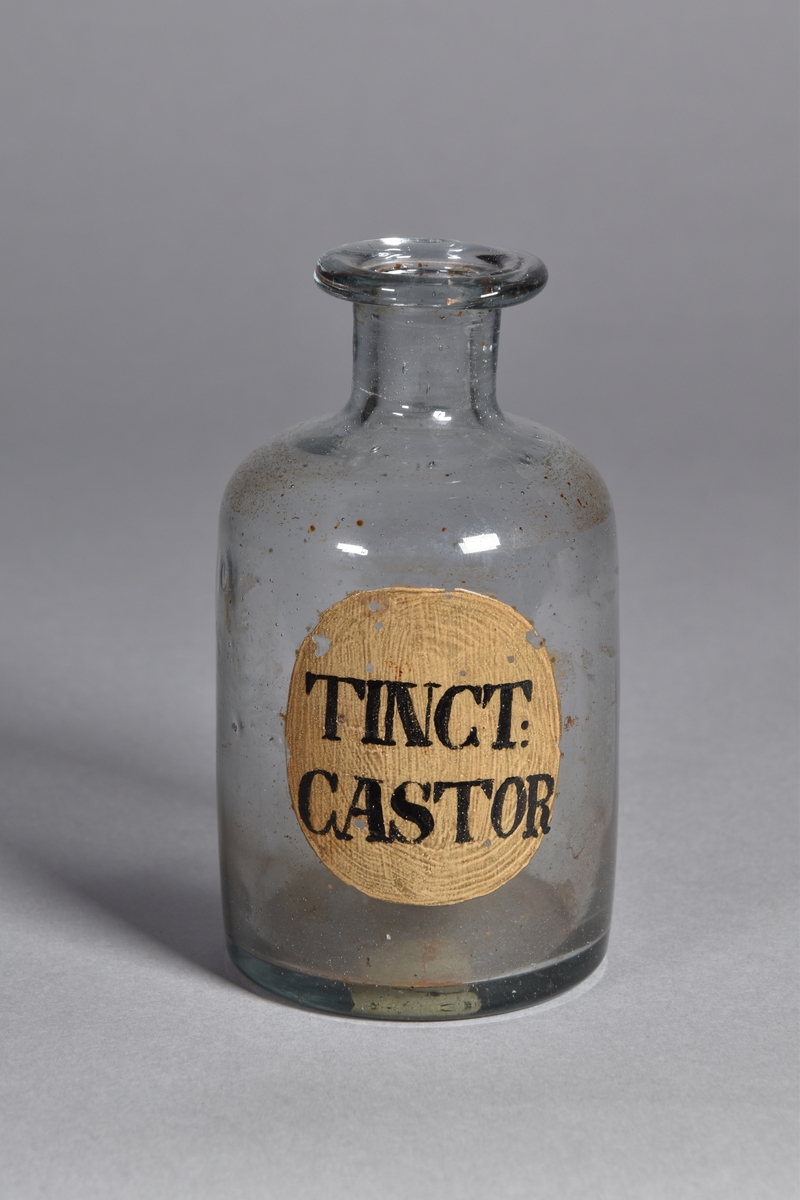Flaska av klart glas, cylindrisk, kort hals med utsvängd mynning. Påmålad etikett med svart text. Smärre rester av innehåll.