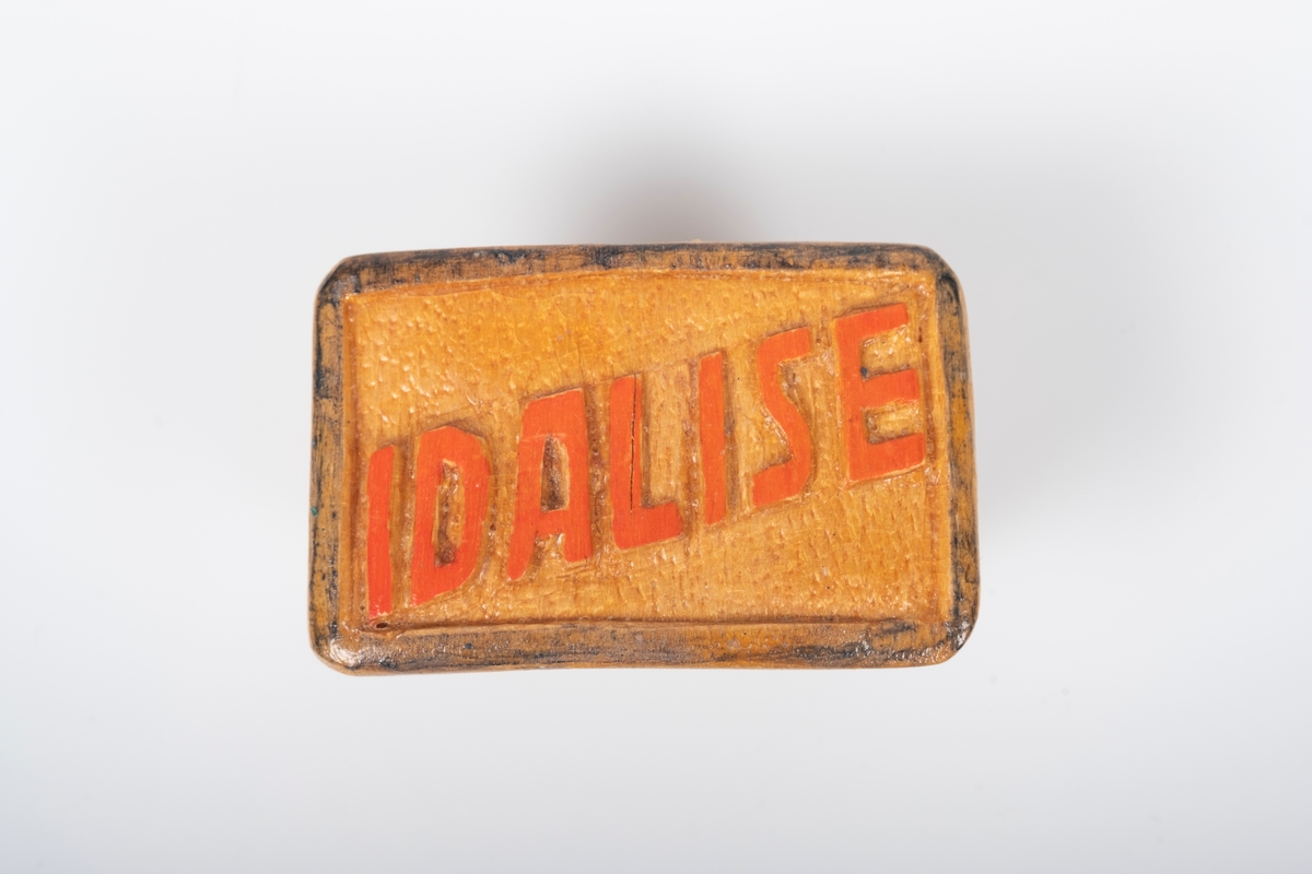 Serviettring laget av tre. På framsiden av serviettringen er navnet "Idalise" skåret ut. Navnet er malt rødt. Det er spikket i buer innvendig, trolig for å holde bedre fast på servietten.