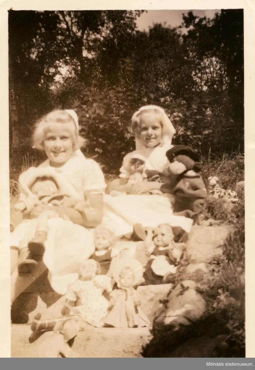Berit Gustafsson och Annemarie Berndtsson sitter i trappan med alla deras dockor och nallen "Jack" i trappan utanför ett av husen i "Stusshålan", Kryssgatan i Mölndal.
1945.