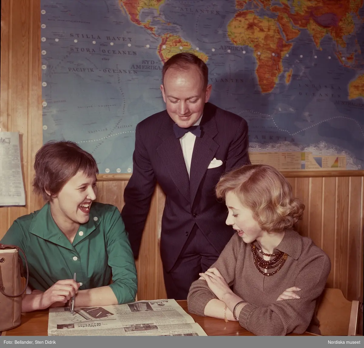 Man och två kvinnor ser på tidning, världskarta i bakgrunden.