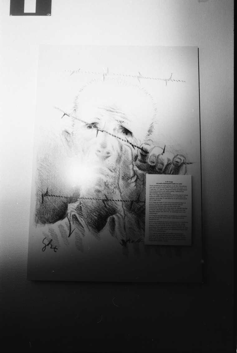 Dokumentation av utställning: G H Trapp och tre sekler av främlingshistoria. Porträtt av koncentrationslägerfånge som kika fram bakom taggtråd, teckning av G H Trapp.