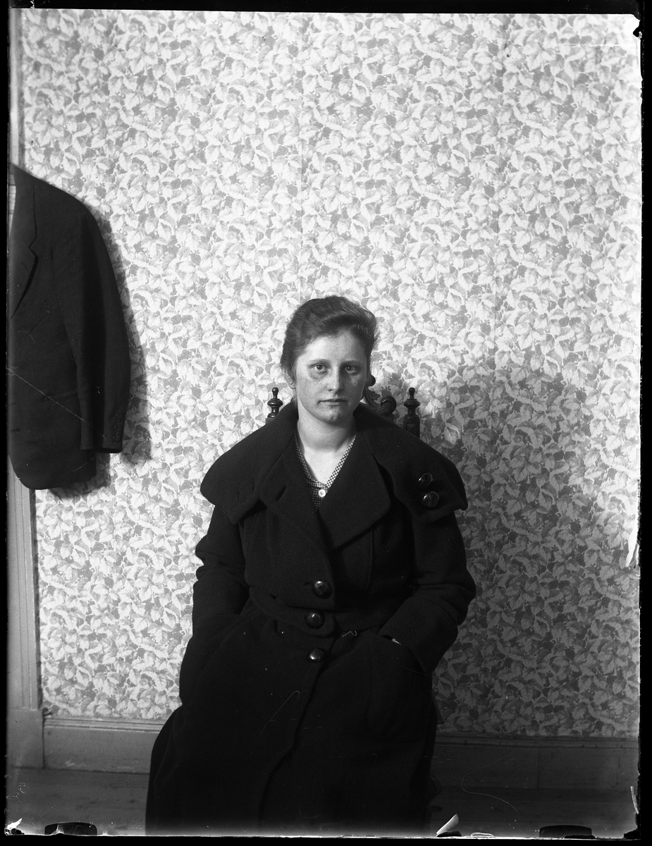 Porträtt av Elise Weber som sitter på en stol i ytterrock och med händerna i fickorna. Bakom henne är en blommig tapet och bredvid henne hänger en mörk kavaj.
