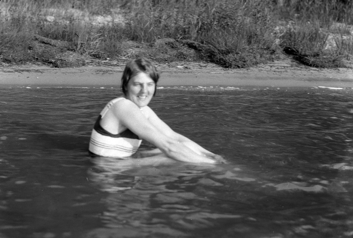 En kvinna, tolkat som Lizzie Skoglund, i baddräkt sitter i vattnet. I fotografens egna anteckningar står det "Besök på Ekholmarna".