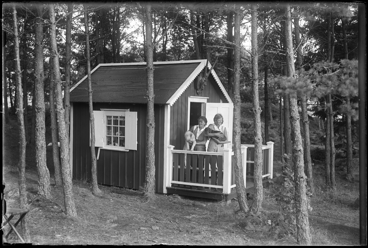 Två kvinnor, med varsin hund i famnen, står på verandan till en liten stuga. I fotografens egna anteckningar står det "Greta Nilssons ö (Stugan)".